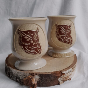 Matera ceramiczne w kształcie kielicha w kolorze kremowym z delikatnym nakrapianym efektem i z rzeźbioną sową w kontrastowym brązowym kolorze umieszczoną w kole.